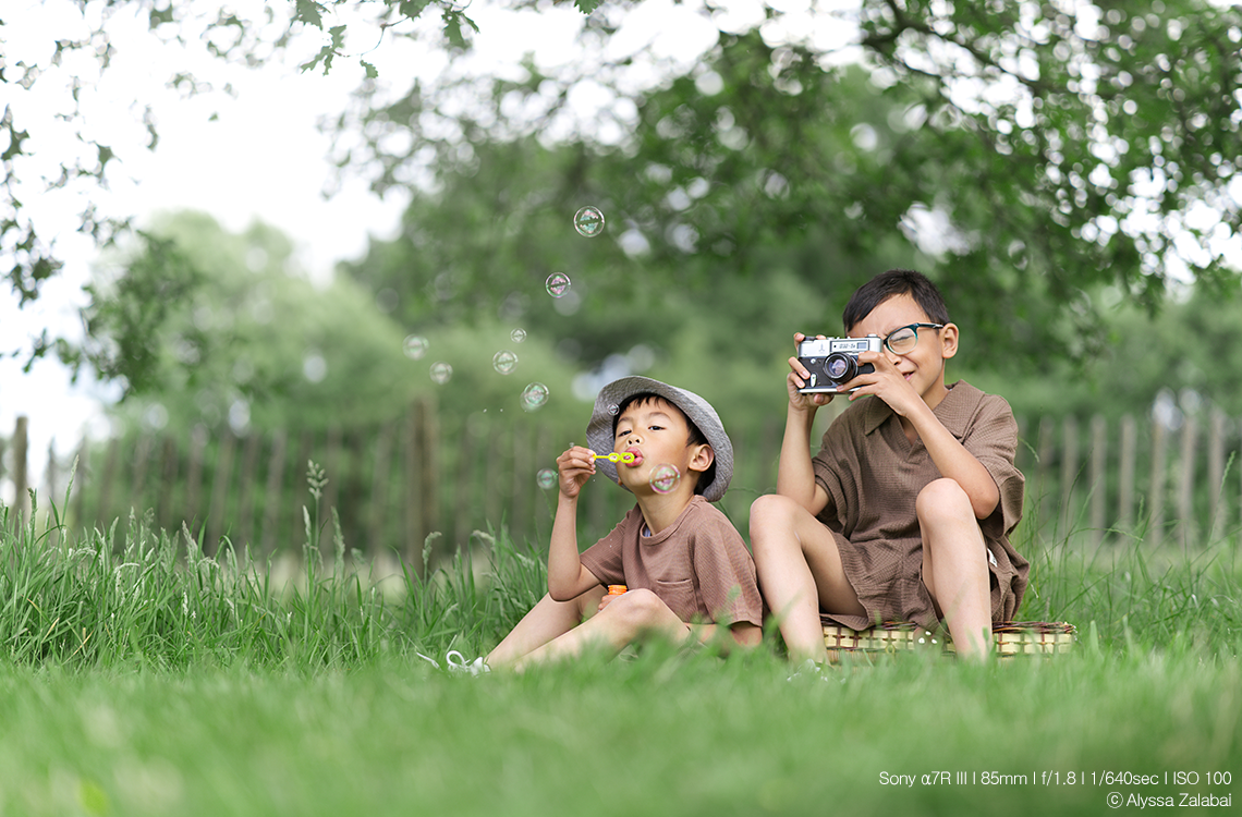 Two children sitting on grass blowing bubbles and taking photos with a camera.